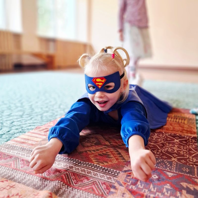 Vaikas su superherojaus kauke ir apsiaustu tupi ant kilimėlio