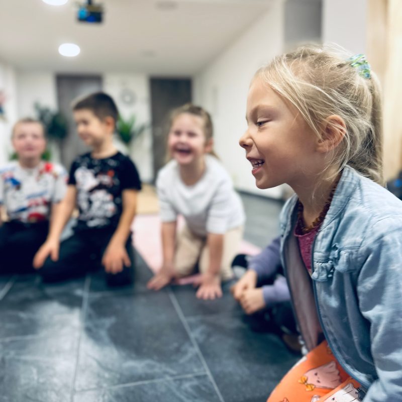 Vaikų grupė sėdi ratu ant grindų ir juokiasi jogos užsiėmimo metu