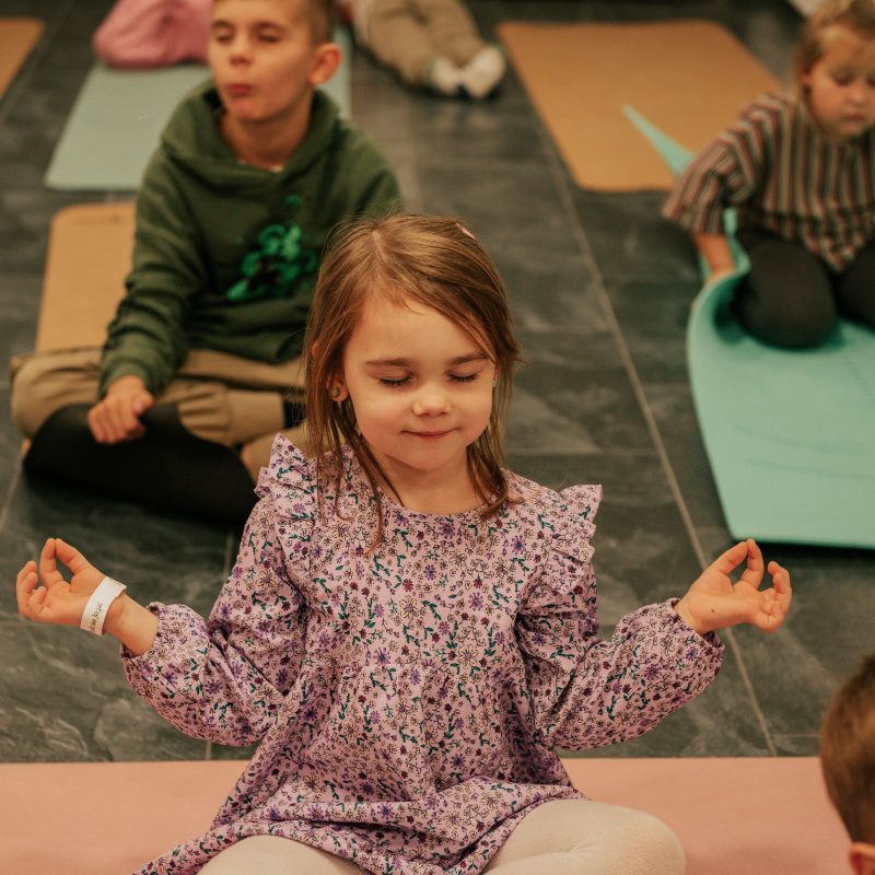Mergaitė sėdi sukryžiavusi kojas ant jogos kilimėlio Pueri Yoga užsiėmime Ukmergėje